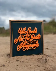 Handgemaakte 3D-geprinte kalligrafie met de tekst “Idle Hands Are The Devil’s Playground” op houten bord met hardhouten lijst