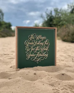 Sierlijke 3D-geprinte kalligrafie met de tekst “The Magic You're Looking For Is In The Work You're Avoiding” op een houten bord met lijst