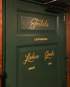 Handgeschilderde gouden scriptbelettering op donkergroene deuren in O' Malley's Irish Pub, uitgevoerd met enamel verf in traditionele signpainting-stijl.